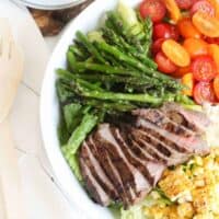 big white bowl of romaine topped with sliced grilled steak, grilled corn and asparagus, and halved tomatoes next to a stack of small white plates