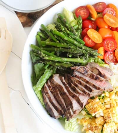 big white bowl of romaine topped with sliced grilled steak, grilled corn and asparagus, and halved tomatoes next to a stack of small white plates