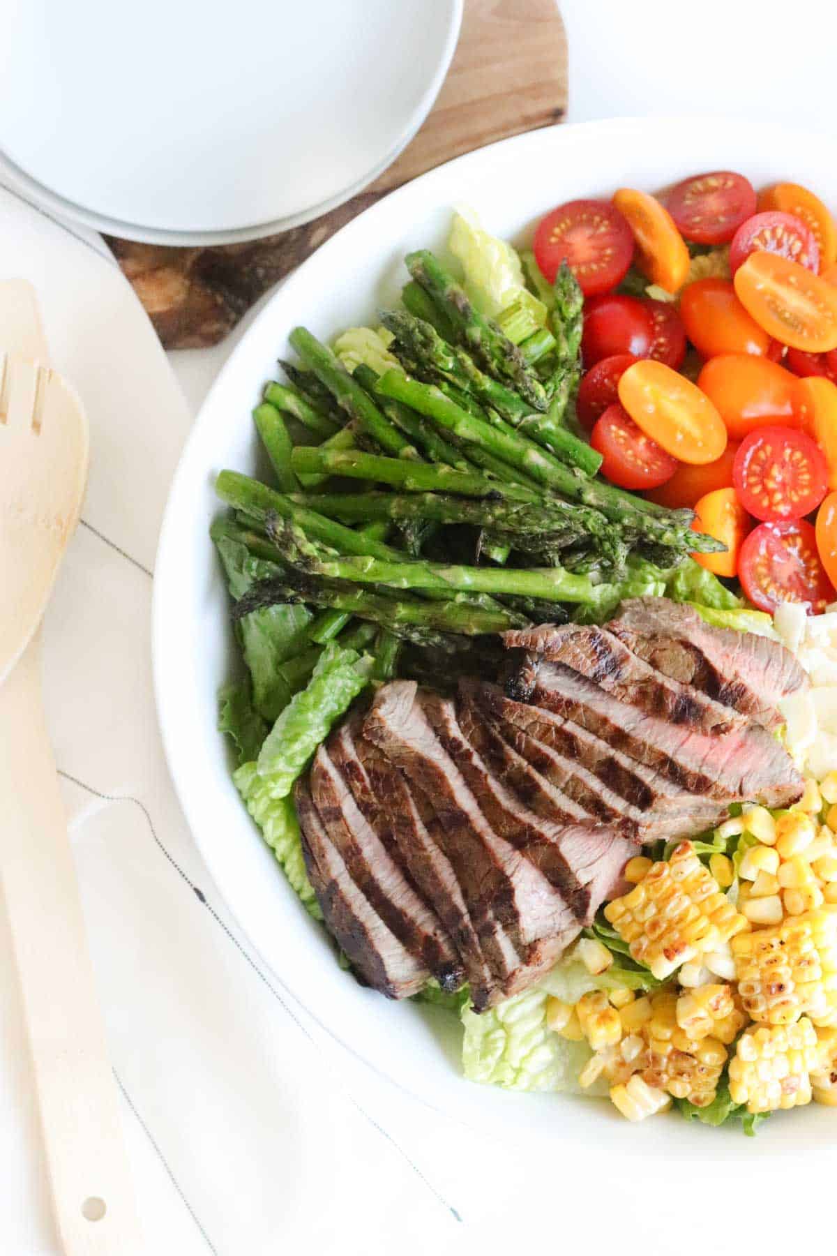 big white bowl of romaine topped with sliced grilled steak, grilled corn and asparagus, and halved tomatoes next to a stack of small white plates