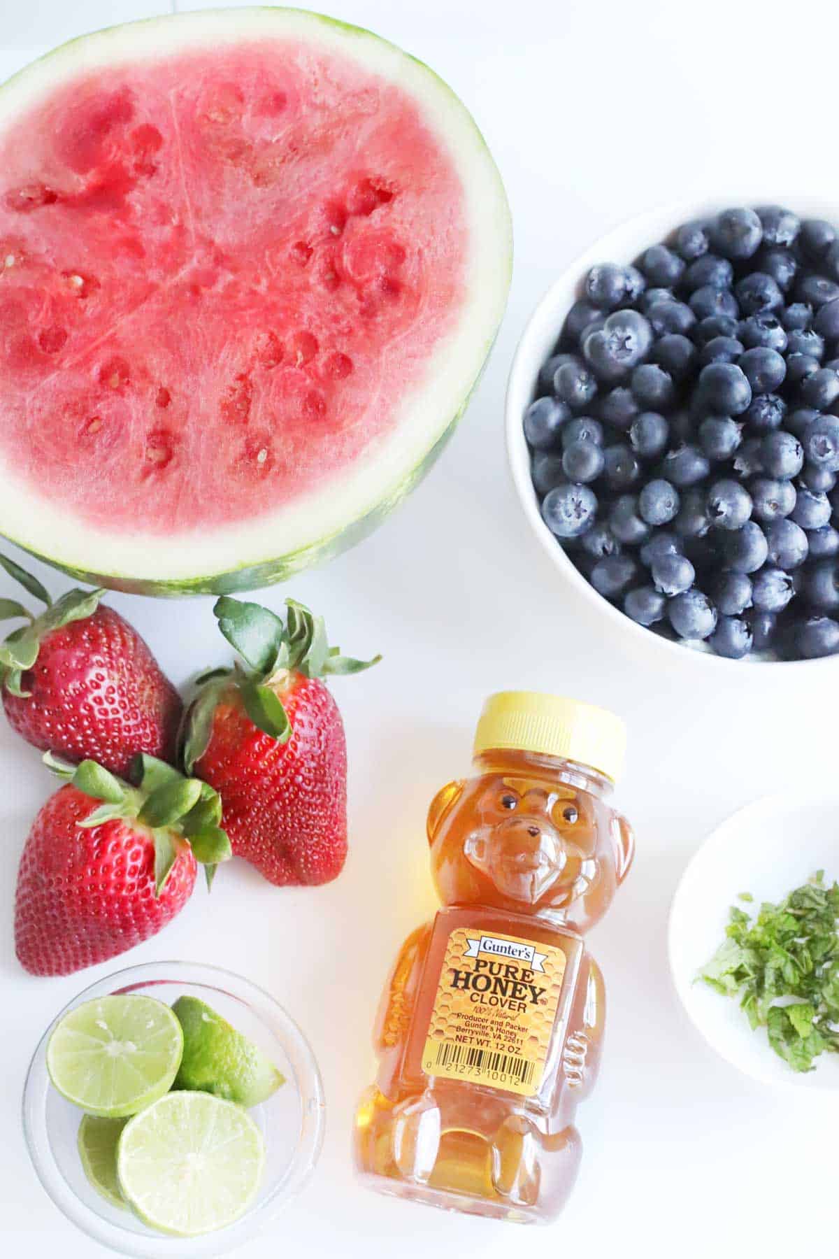 watermelon slice, strawberries, bowls of blueberries, mint leaves and limes, and a bear-shaped bottle of honey