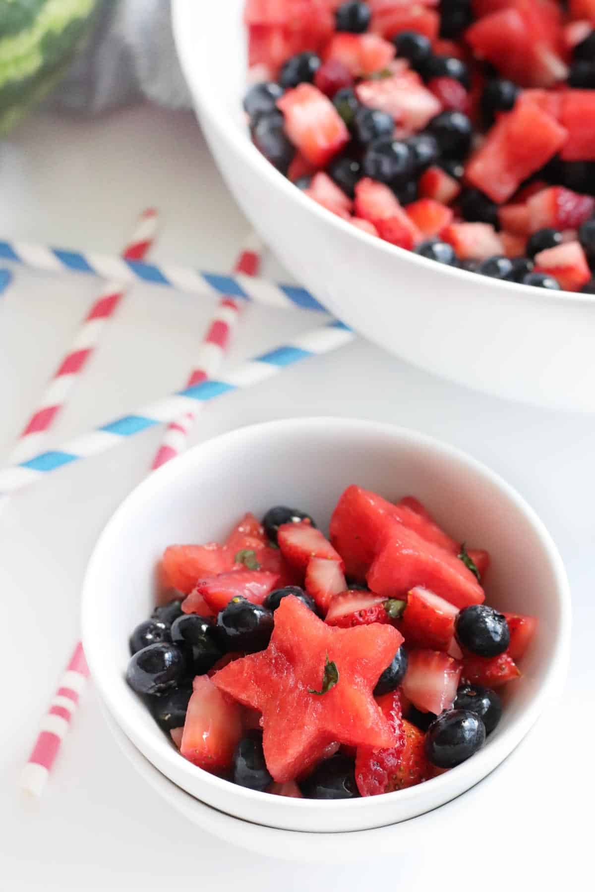 small bowl of watermelon berry fruit salad with a serving bowl and red, white, and blue straws in the background