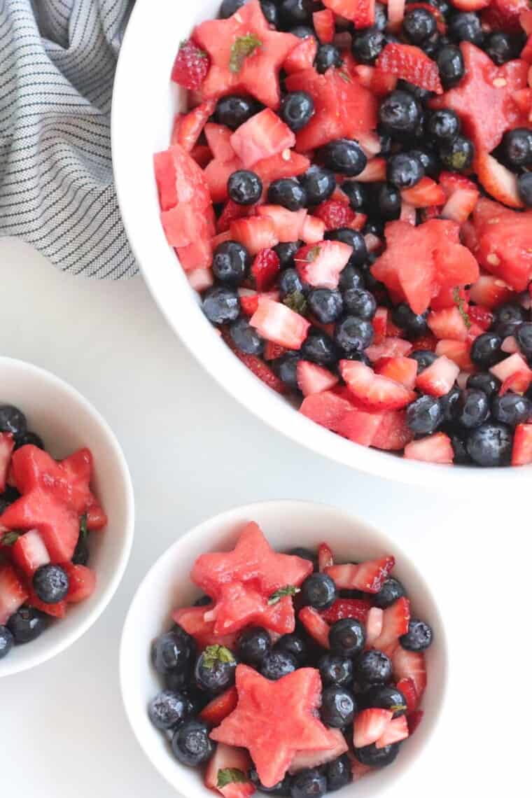 a serving bowl and two small bowls of fruit salad coated in a dressing with mint leaves