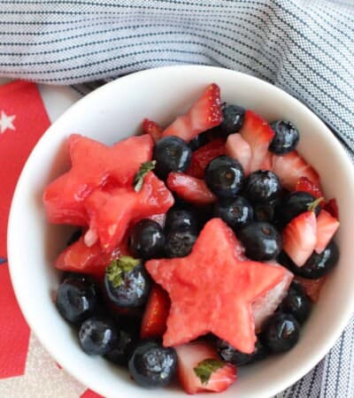 small bowl of fruit salad with chopped strawberries, blueberries, and watermelon stars surrounded by red ad white star napkins and a blue stripe cloth napkin