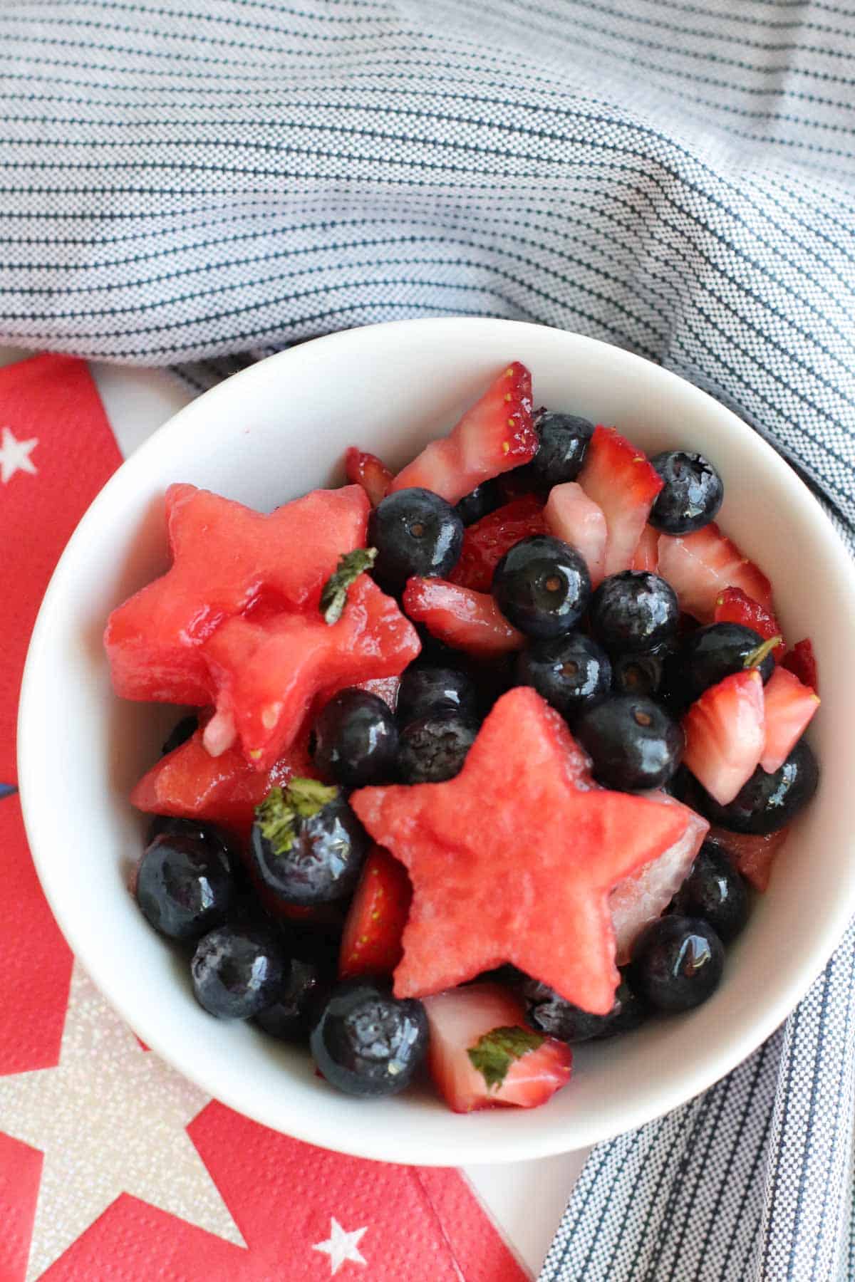 small bowl of fruit salad with chopped strawberries, blueberries, and watermelon stars surrounded by red ad white star napkins and a blue stripe cloth napkin