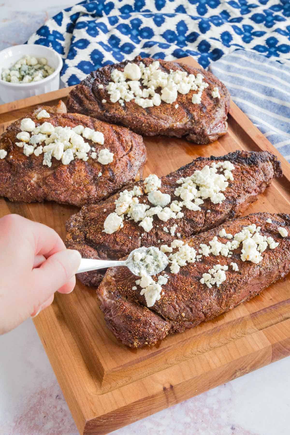 Crumbled Bleu Cheese Being Placed on Top of Blackened NY Strip Steaks
