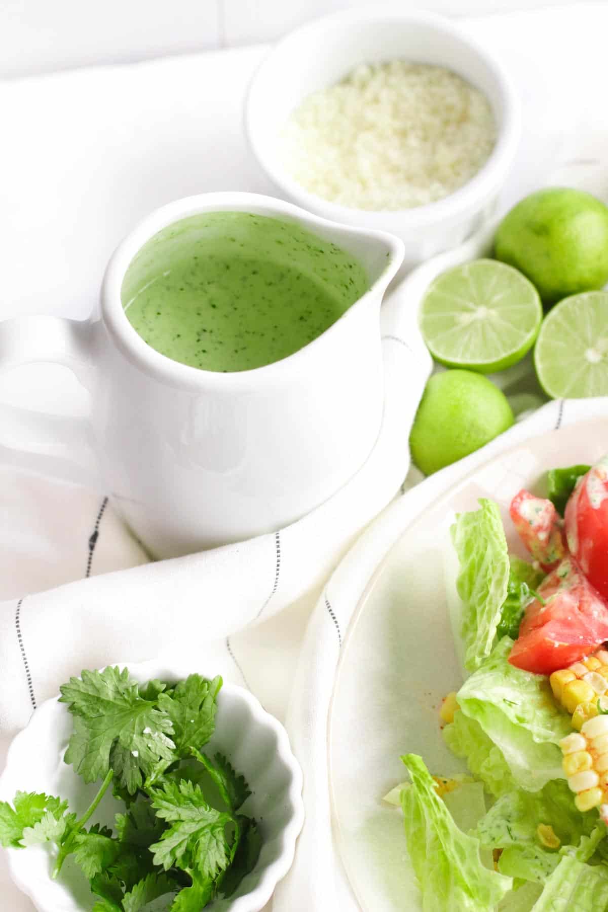 container of dressing, bowls of salad and cilantro, and lime nestled in a napkin on a table