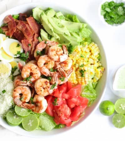 grilled shrimp cobb salad in a serving bowl with lime halves, a container of salad dressing, and a bowl of cheese next to it