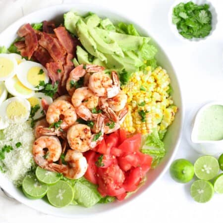 grilled shrimp cobb salad in a serving bowl with lime halves, a container of salad dressing, and a bowl of cheese next to it