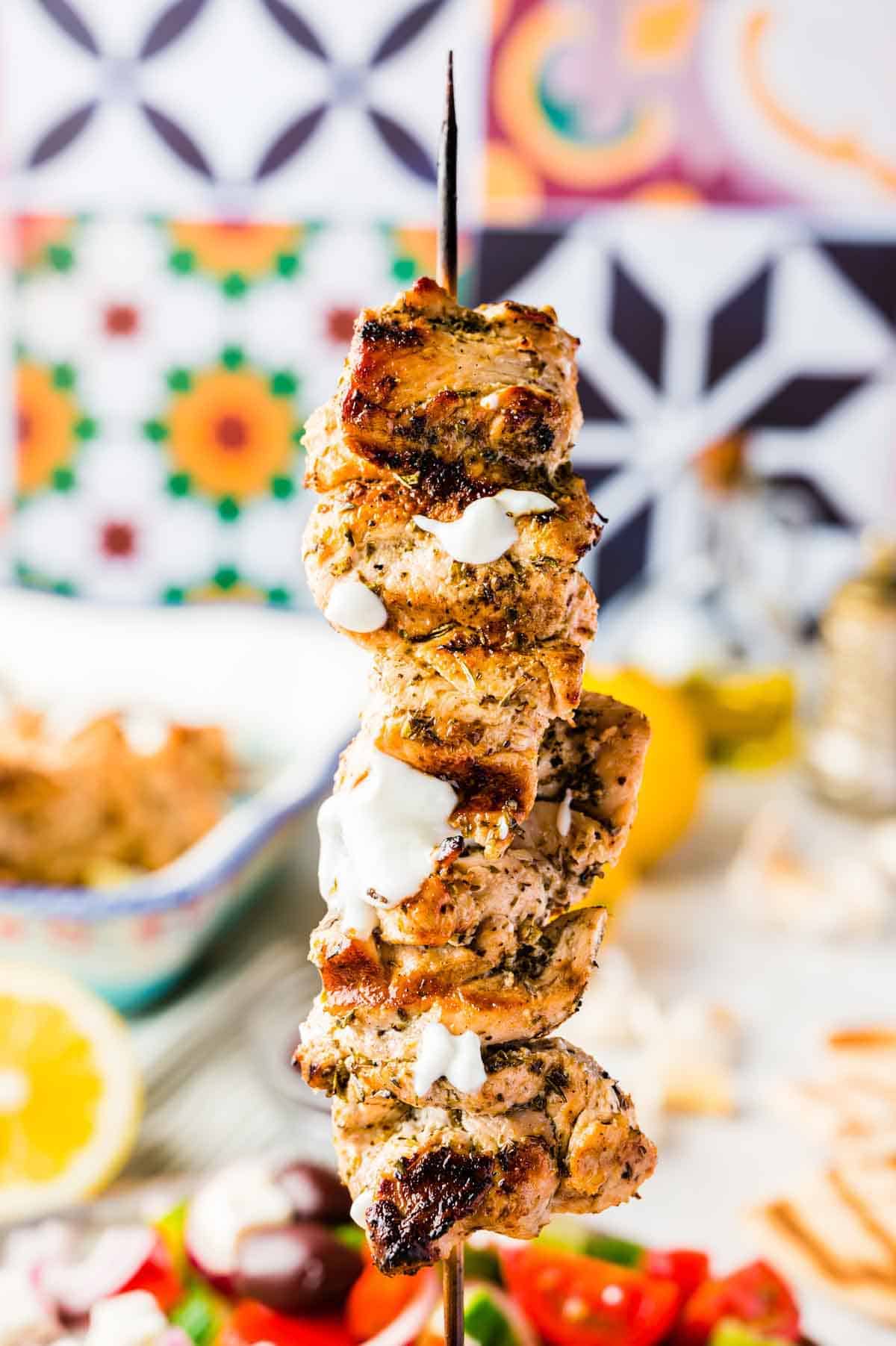 A Single Greek Chicken Skewer with Tzatziki Sauce Being Held in Front of a Colorful Wall