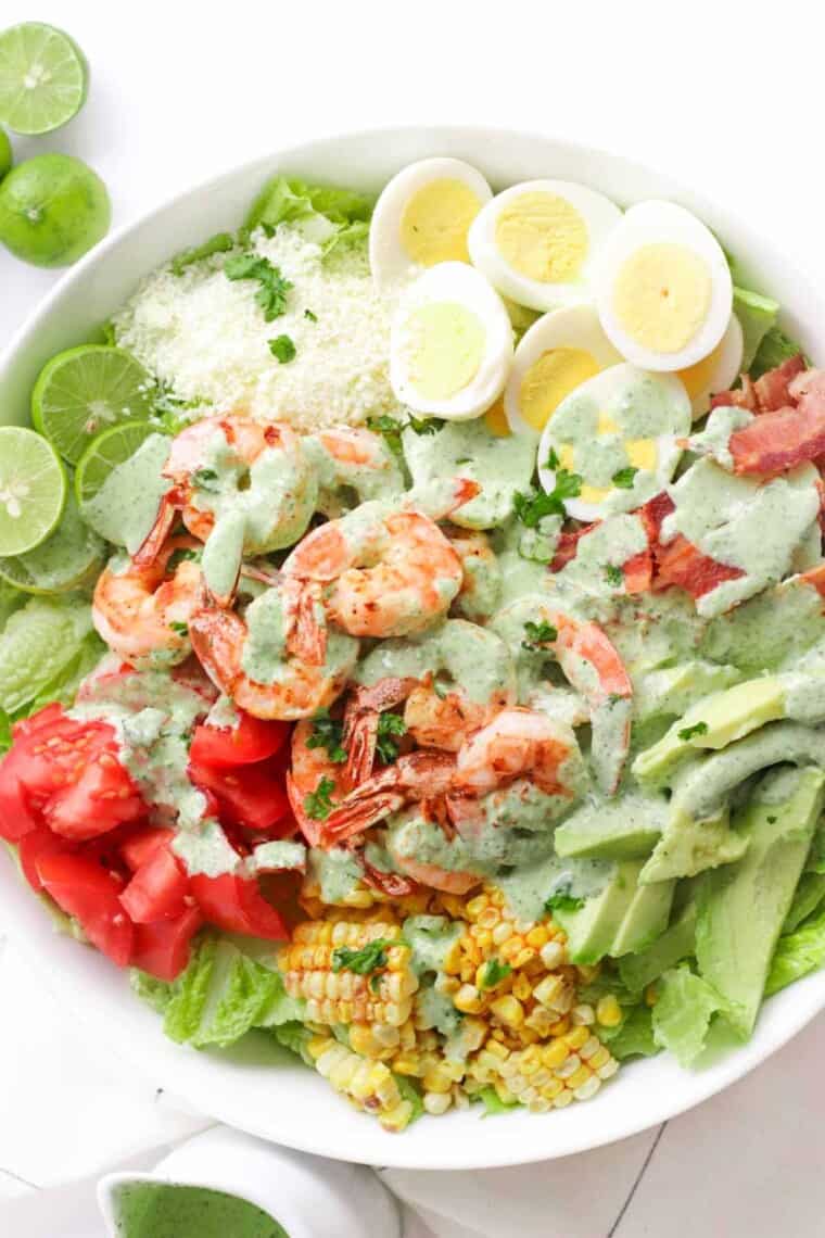 Shrimp Cobb Salad with hard boiled eggs, bacon, limes, tomatoes, avocado, cotija cheese, grilled corn, and cilantro lime dressing in a white serving bowl