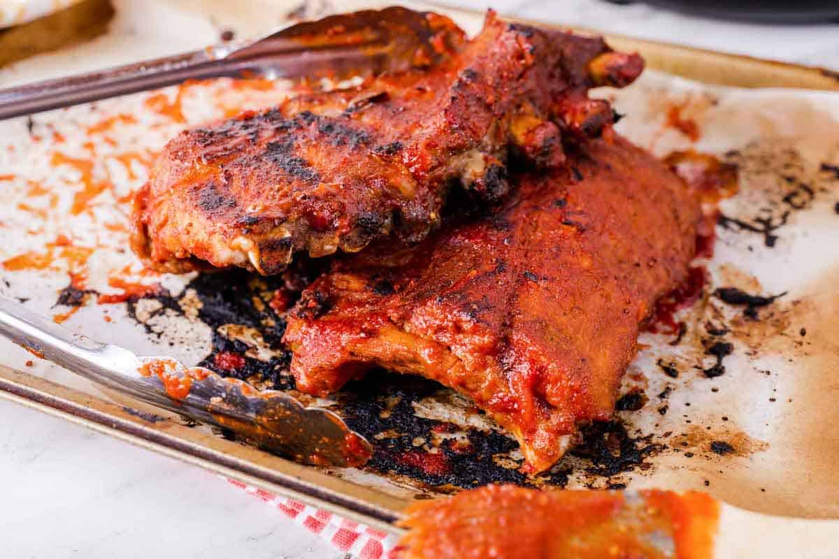 two racks of ribs on a parchment-lined baking sheet with tongs