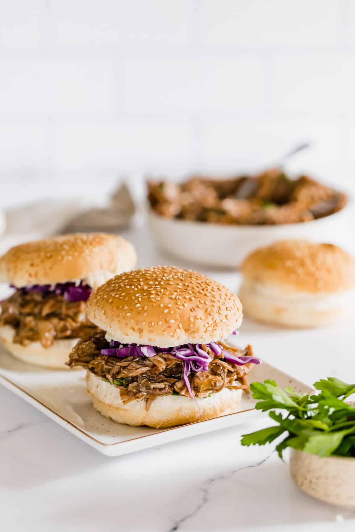 A Platter Holding Two Pulled Pork Sandwiches with a Bowl of Pulled Pork and a Bun in the Background
