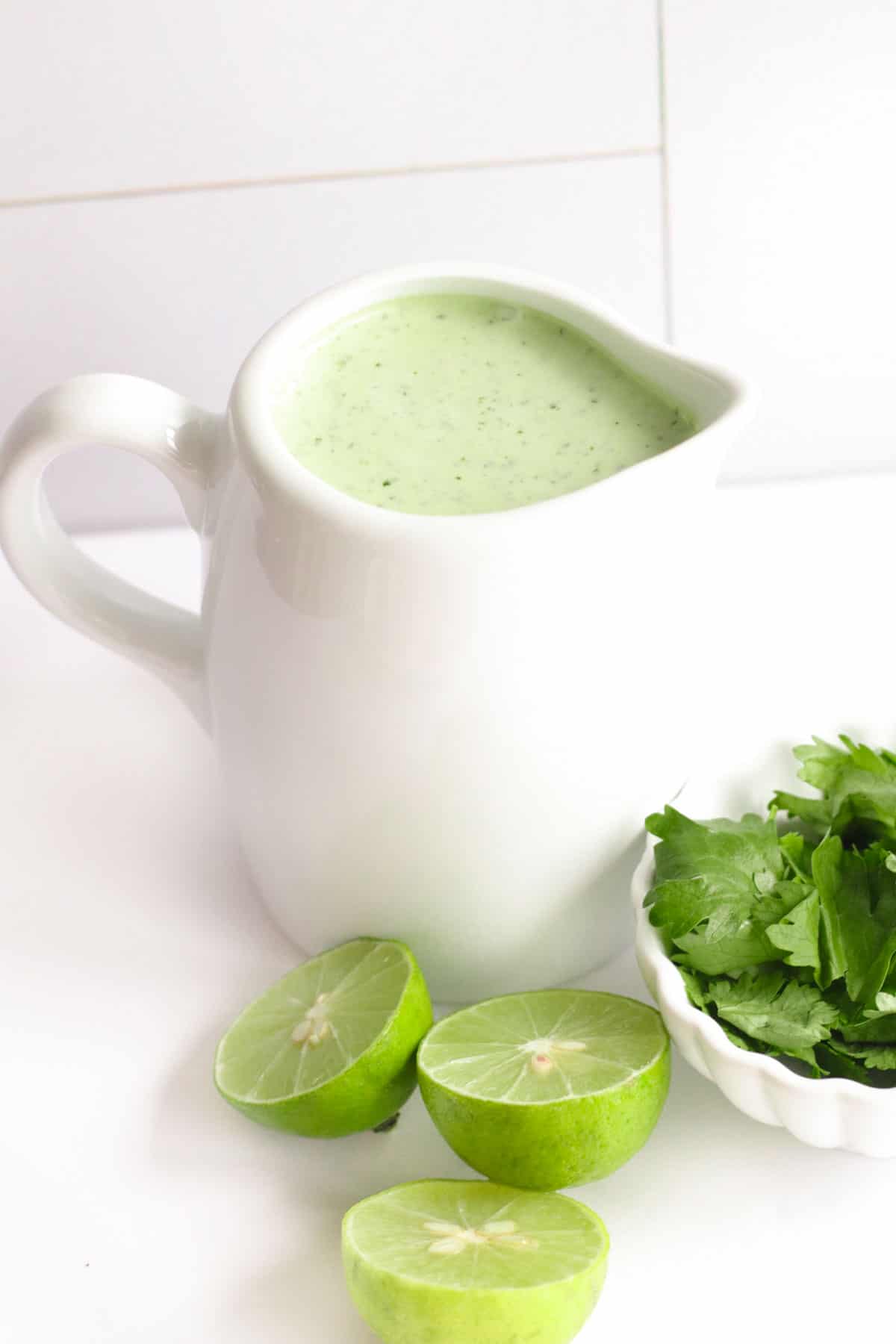 small white pitcher of cilantro lime dressing with limes and cilantro on the table