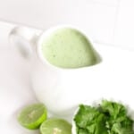 a container of cilantro lime jalapeno ranch salad dressing with a bowl of cilantro and lime halves next to it on a table