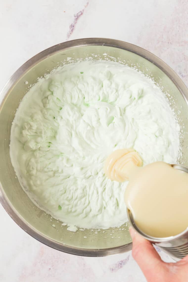 pouring can of sweetened condensed milk into a bowl with the whipped cream tinted green