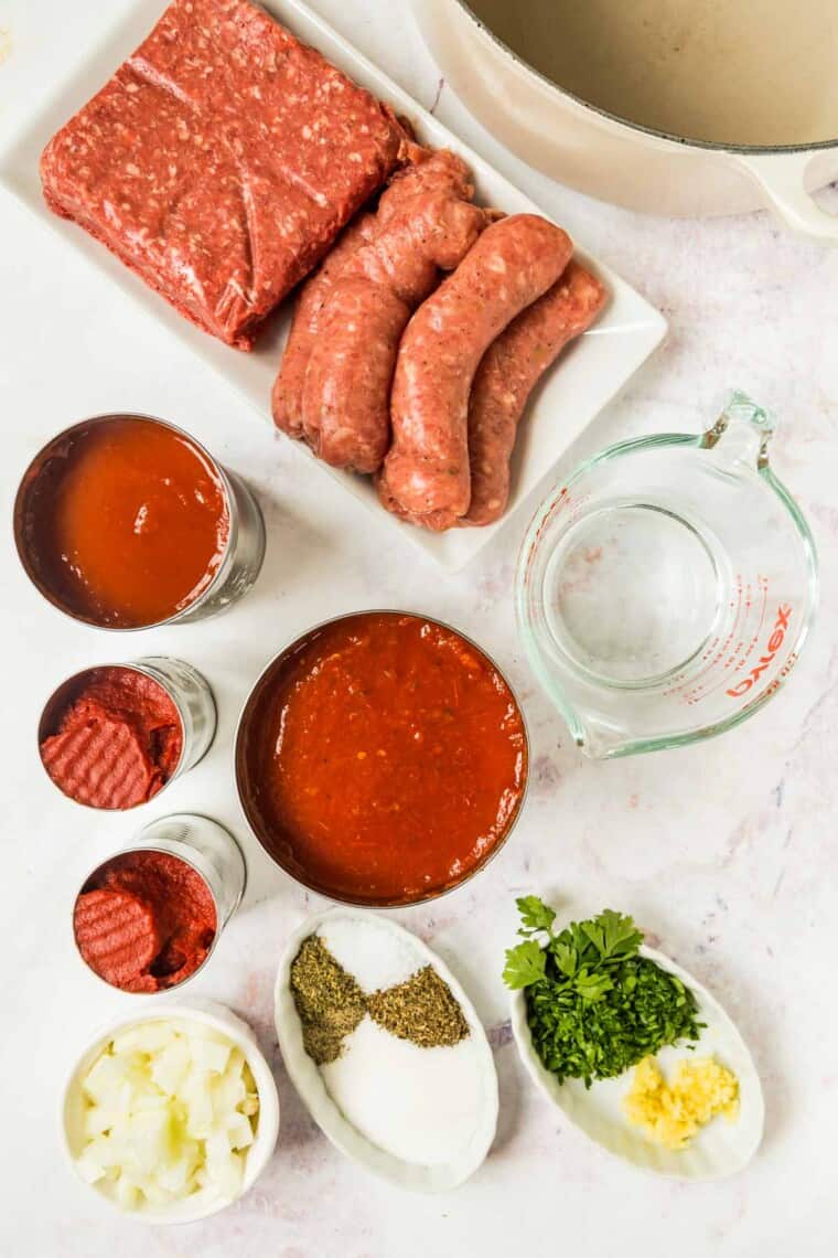 all of the ingredients to make meat sauce for lasagna, including cans of crushed tomatoes and tomatoes paste, chopped onions, garlic, and parsley, raw ground beef and sausage, and dries seasonings
