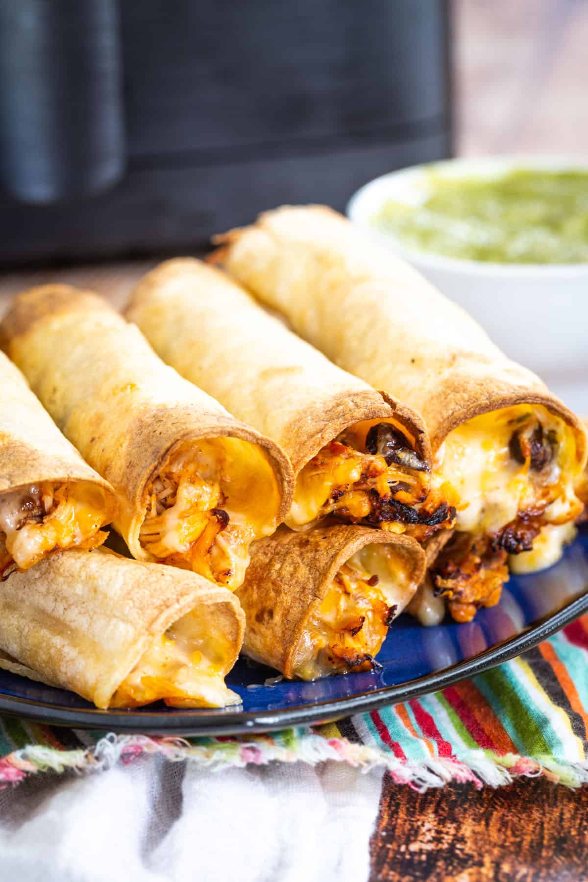 cheesy chicken taquitos stacked on a bleu plate in front of an air fryer and a small bowl of guacamole