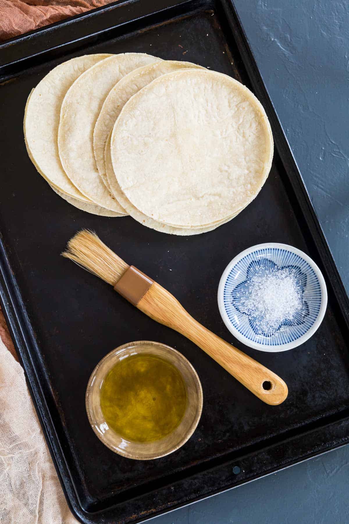 baking sheet with corn tortillas, olive oil, a pastry brush, and a small bowl of kosher salt