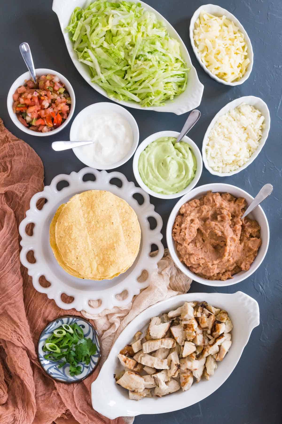 a plate of tostada shells with bowls of fully cooked chicken strips, lettuce, crumble cotija cheese, refried beans, salsa, sour cream, guacamole, and cilantro and scallions