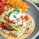 a chicken tostada with refried beans, lettuce, salsa, cheese, and sour cream served on a blue plate with mexican rice