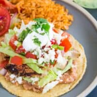 a chicken tostada with refried beans, lettuce, salsa, cheese, and sour cream served on a blue plate with mexican rice