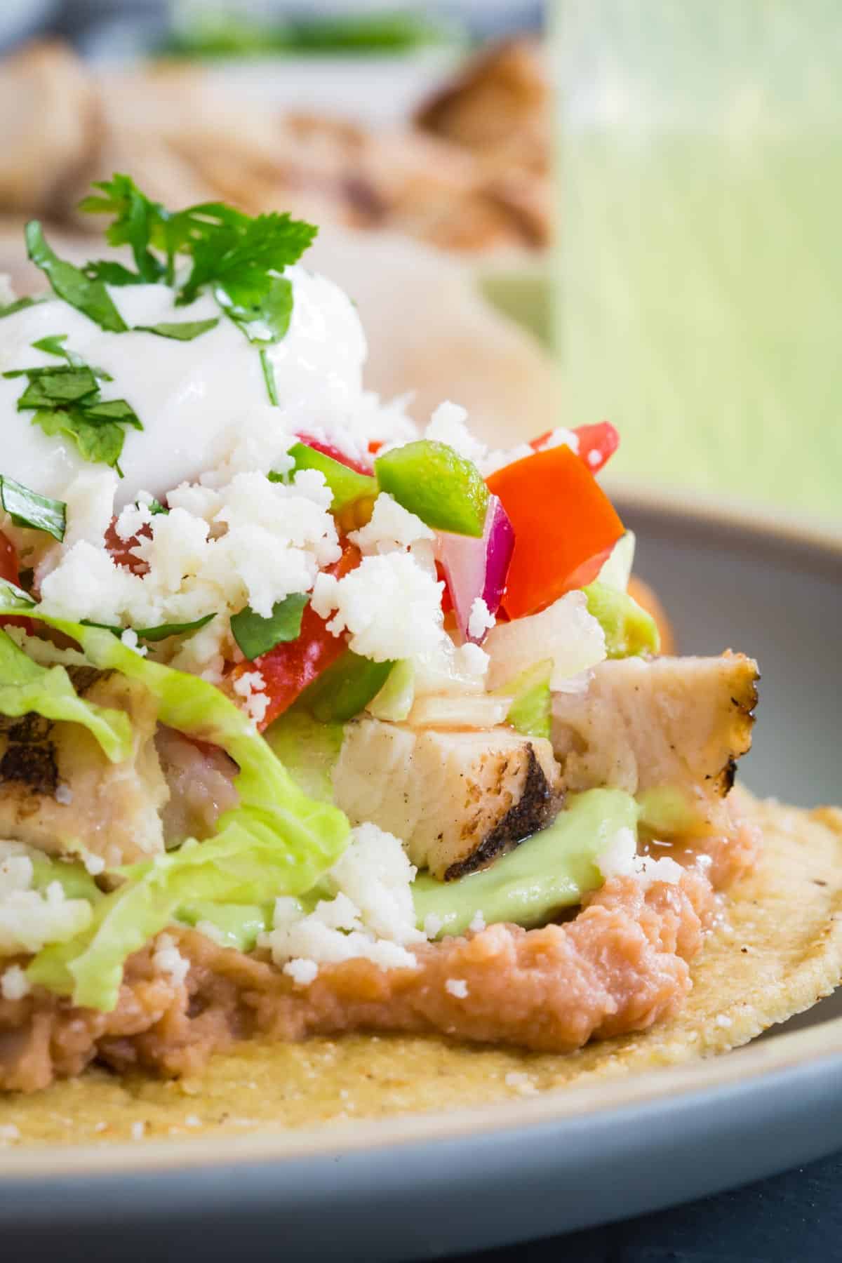 a crispy corn tortilla topped with layers of refried bans, guacamole, cooked chicken pieces, lettuce, salsa, crumbled cotija cheese, sour cream and a garnish of cilantro