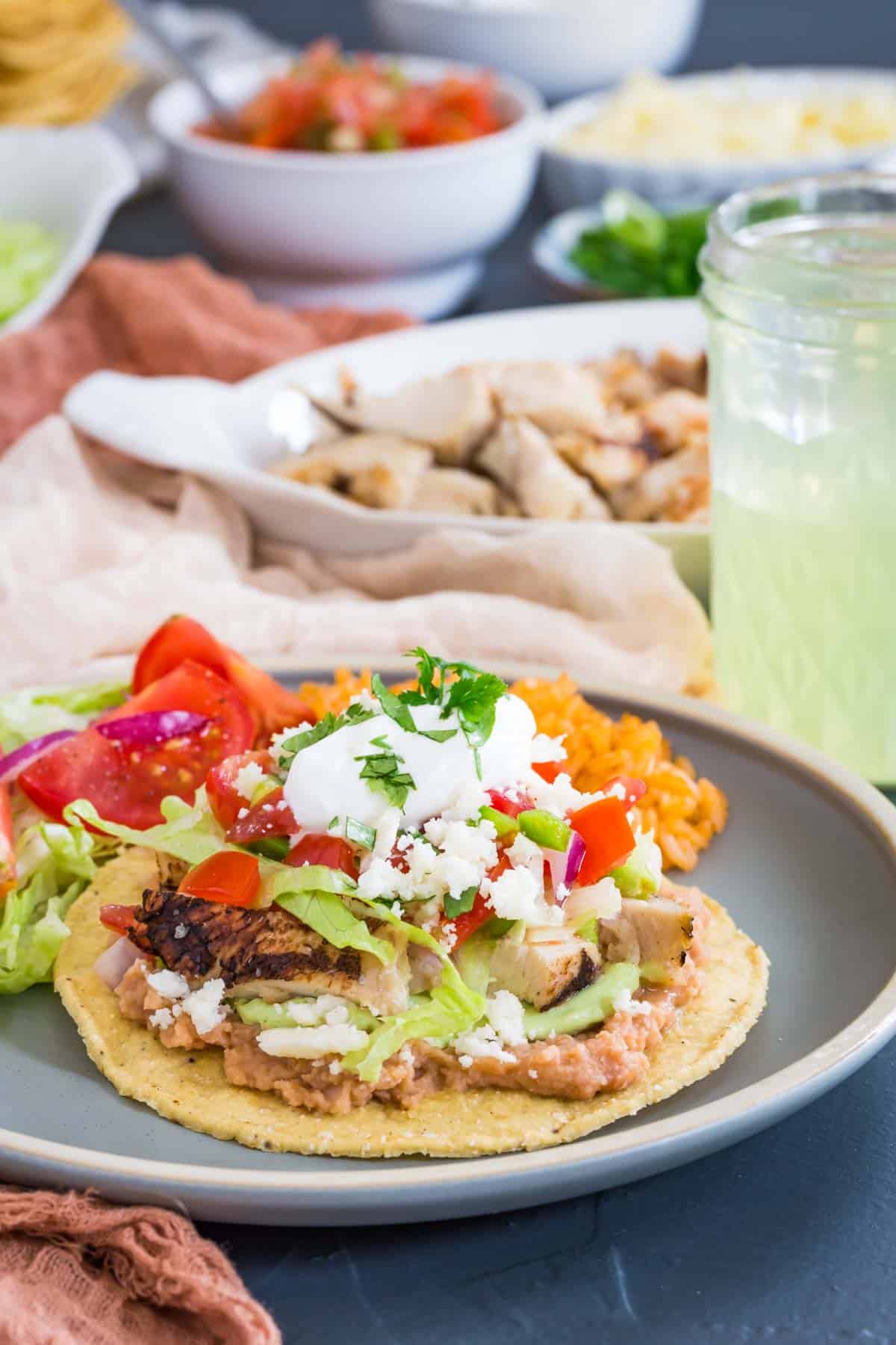 a chicken tostada on a plate with rice and bowls of all of the toppings in the background