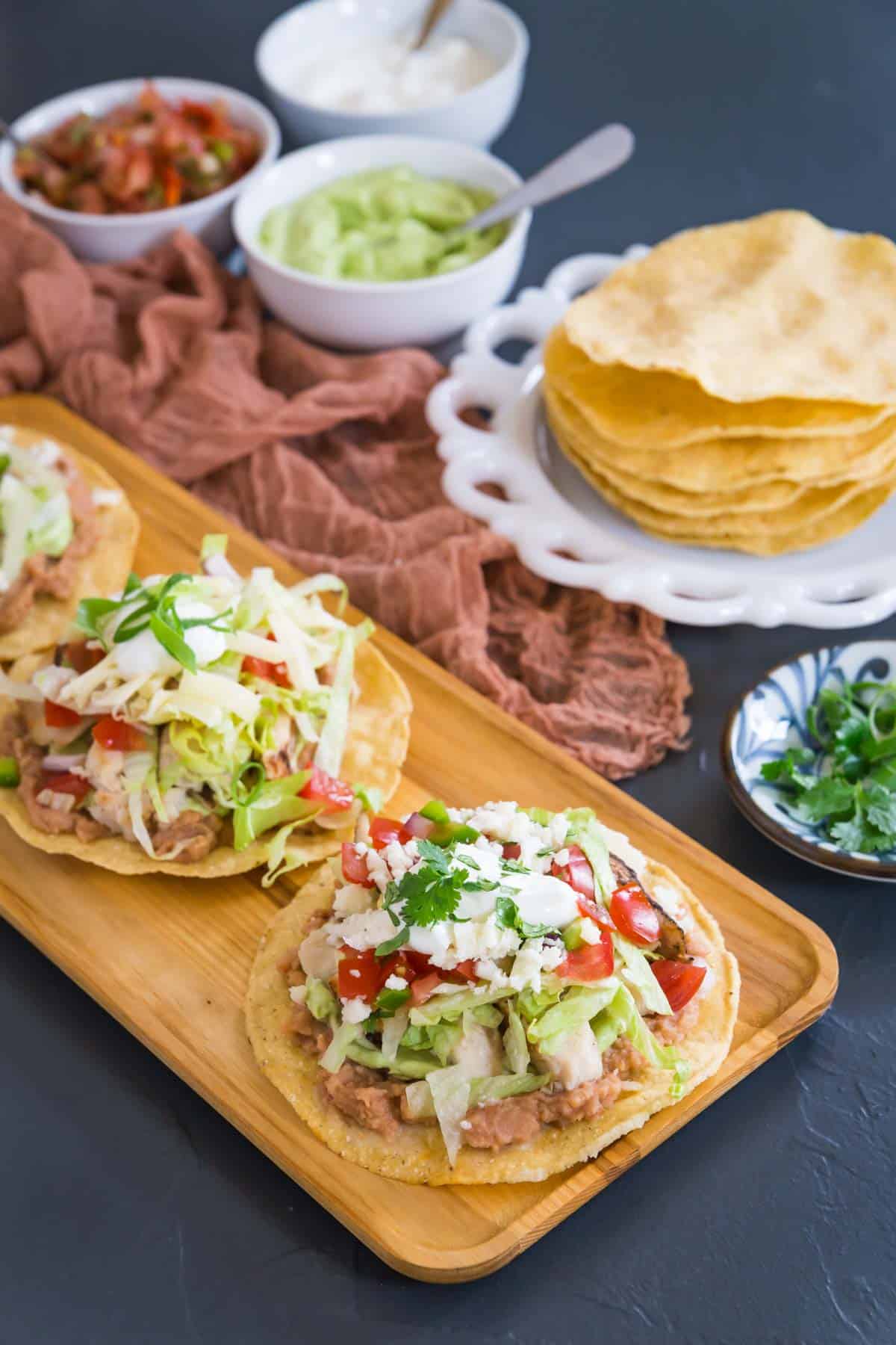 three chicken tostadas on a wooden serving platter with a plate of tostada shells and bowls of salsa, sour cream, and guacamole