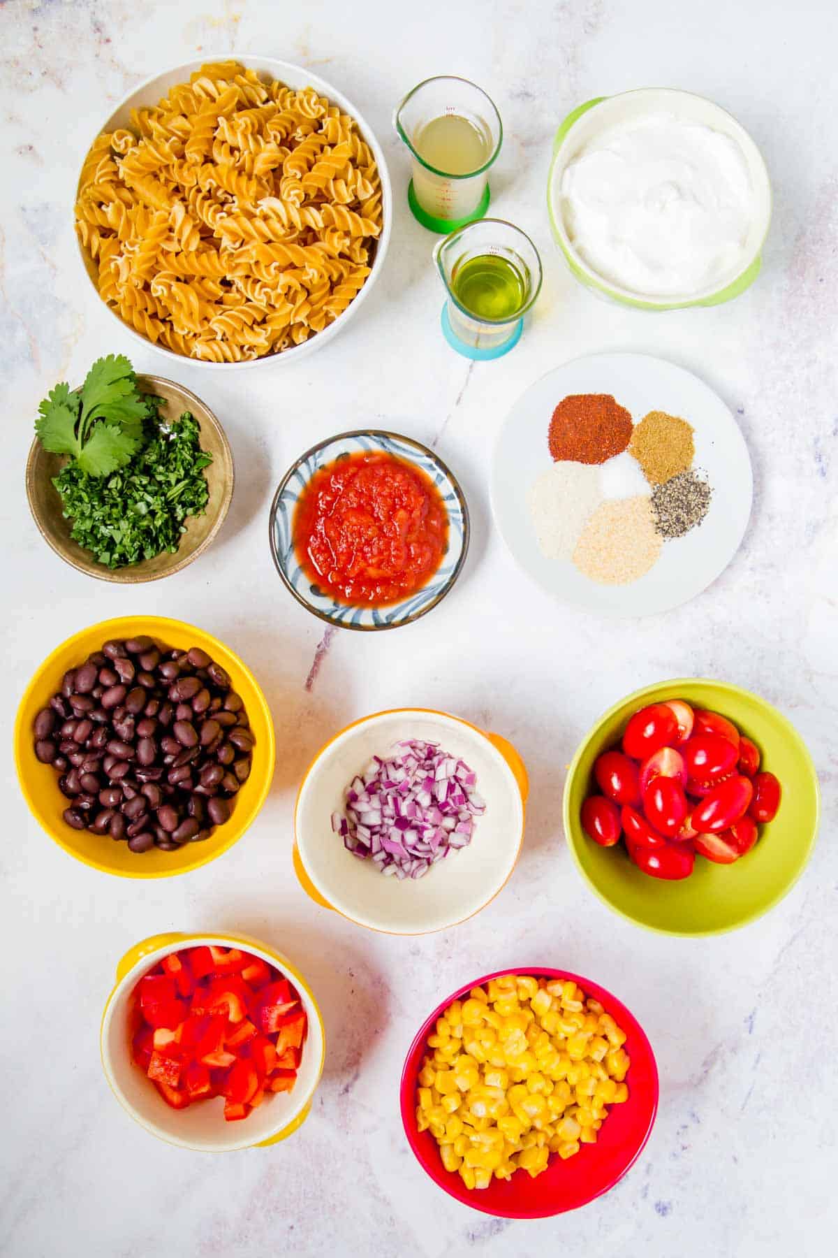 bowls of uncooked pasta, corn, black beans, chopped red pepper, halved grape tomatoes, minced red onion, greek yogurt, salsa, and cilantro, olive oil and vinegar in small measuring beakers, and a plate with spices