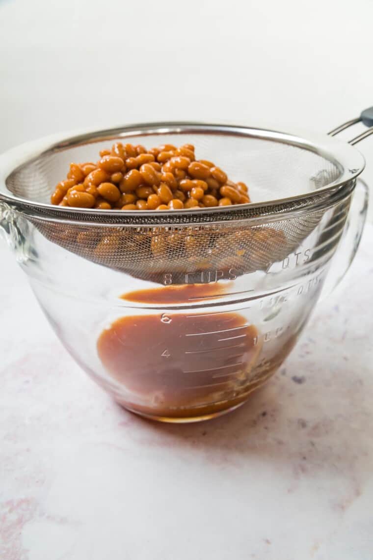 canned baked beans in a mesh strainer over a glass bowl with the liquid collecting in the bowl