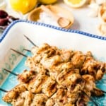 Greek chicken kabobs on wooden skewers in a serving platter surrounded by lemons, garlic cloves, and other condiments