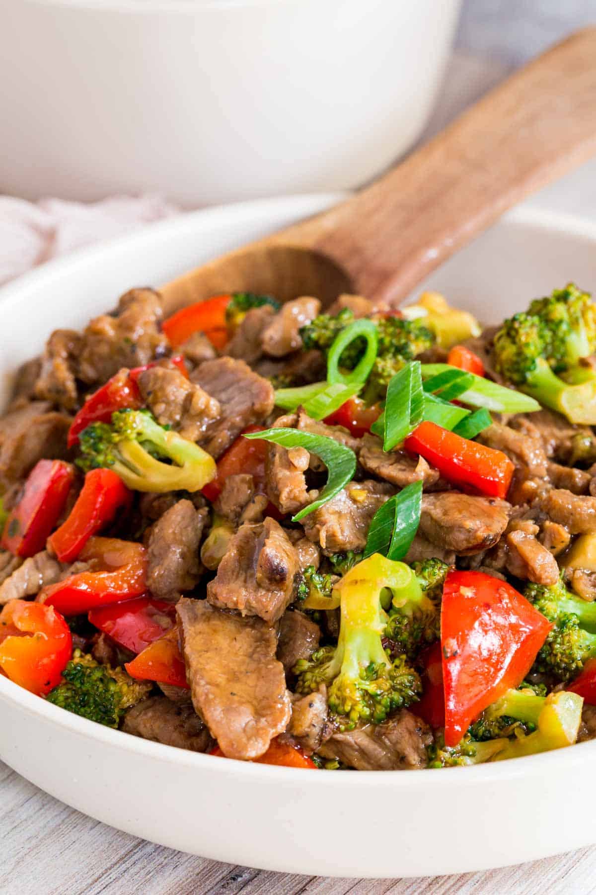 stir fry of steak strips, broccoli and red peppers in a sauce topped with sliced scallions in a white serving bowl