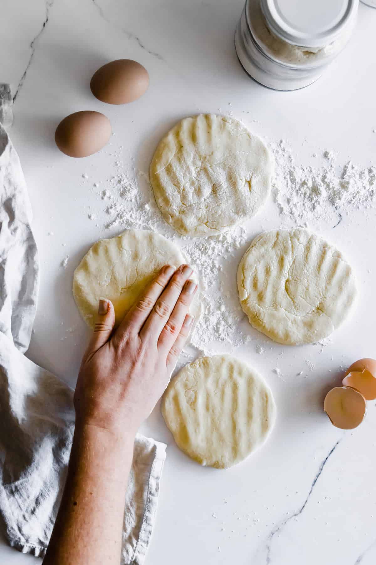 Four Discs of Gluten Free Pita Dough on a Marble Countertop with a Hand Pressing Down on One of Them