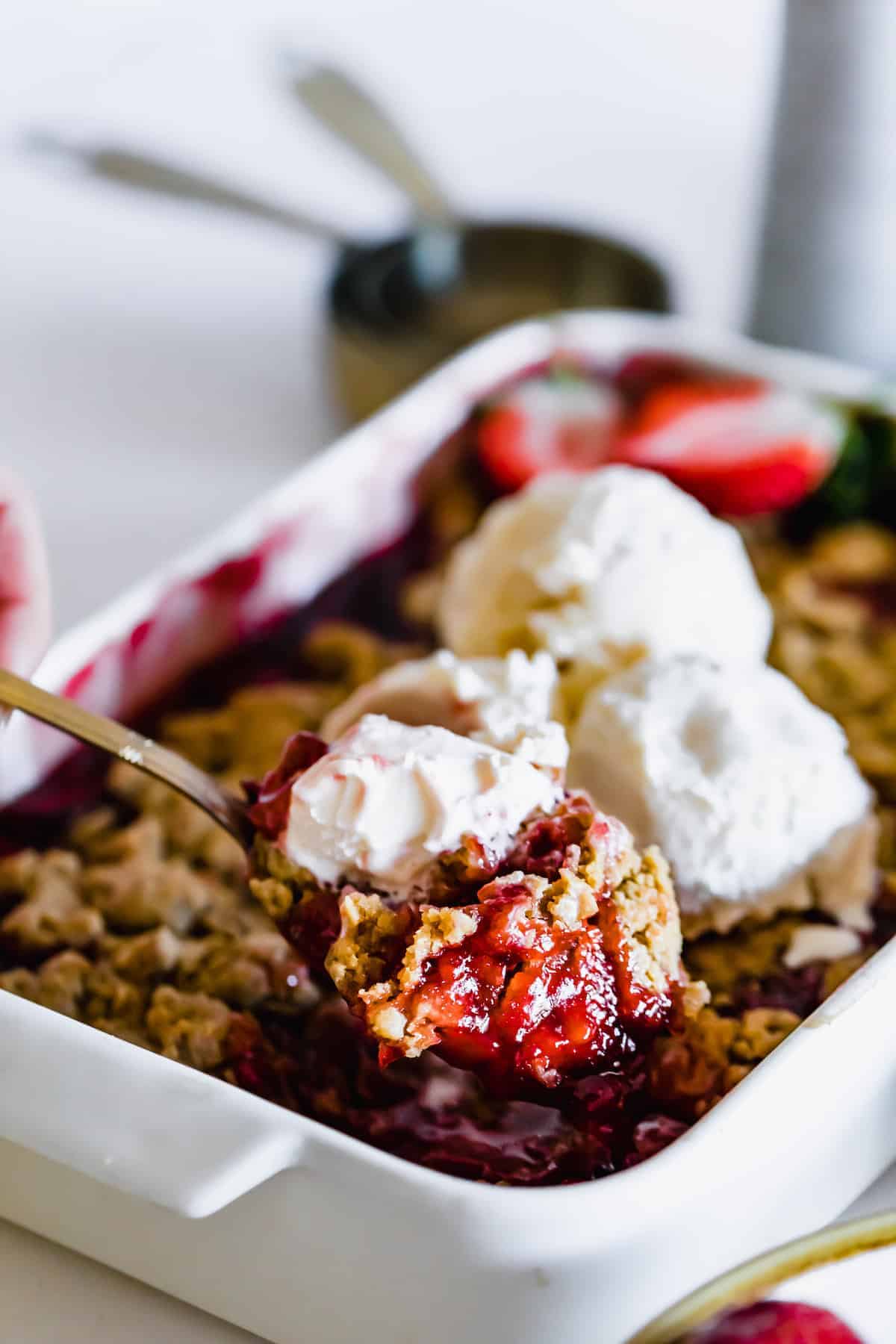 A Metal Spoon Holding a Scoop of Strawberry Crumble Over a Pan Full of the Dessert