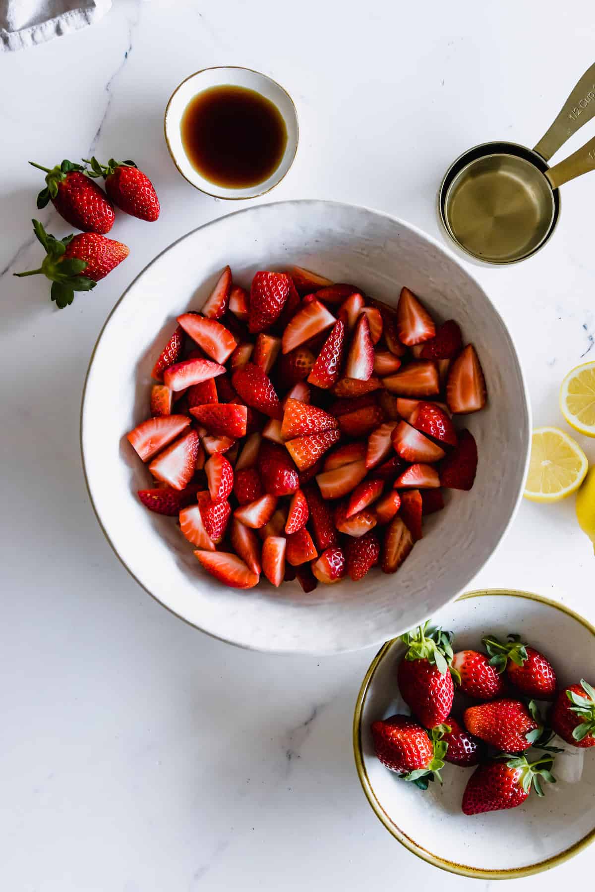 A bowl of fresh strawberries cut into quarters beside a dish of vanilla extract and two measuring cups.