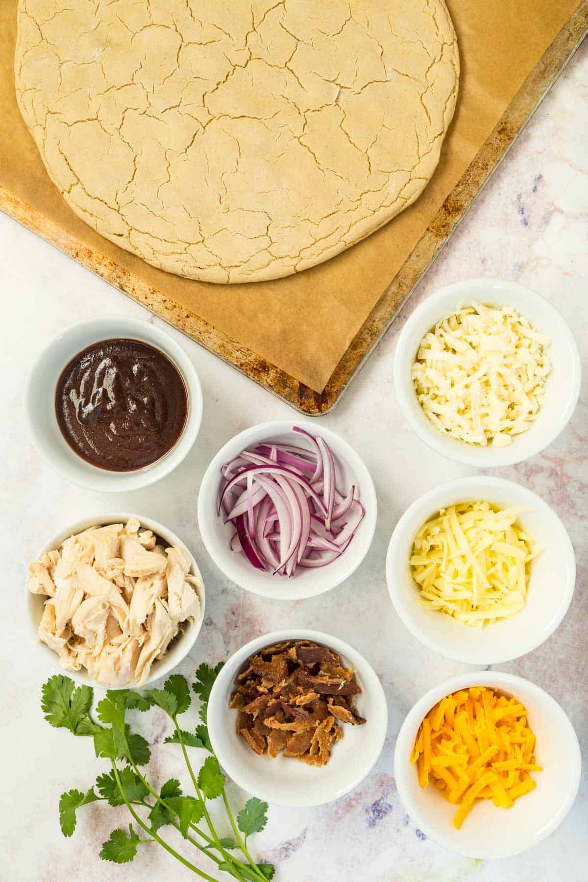 pizza crust plus bowls of barbecue sauce, chicken, three kinds of cheese, crumbled bacon, and sliced red onions, plus a bunch of cilantro on a marble tabletop