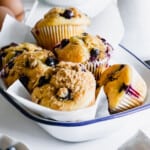 six blueberry muffins piled in a white bowl lined with wax paper