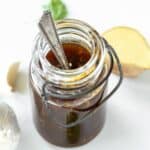 teriyaki sauce in a glass jar with a spoon in it next to a piece of ginger and a bulb of garlic