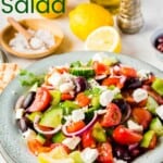 serving bowl of Greek salad with tomatoes, peppers, olives, cucumbers, onions, and feta on a table with lemons, olive oil, and salt