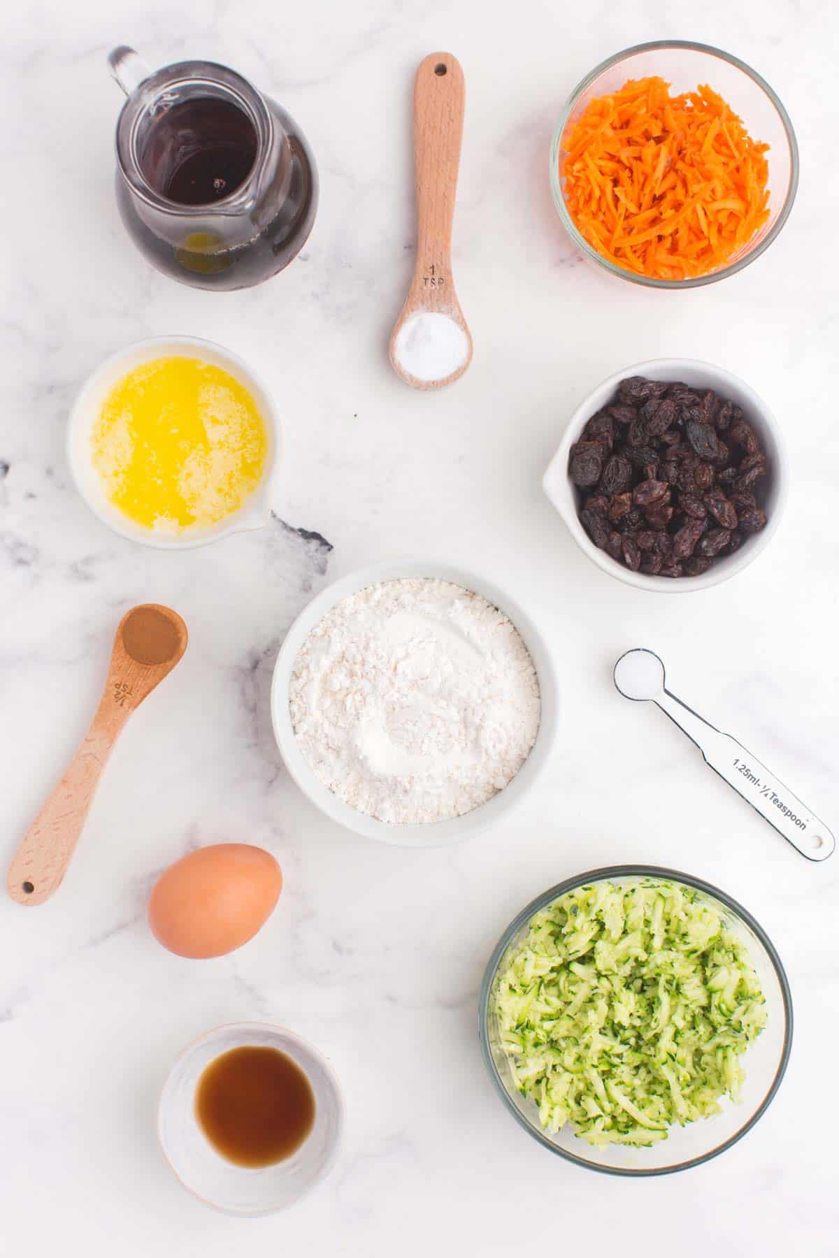 bowls of flour, shredded carrots and zucchini, vanilla extract, raisins, and melted butter, a pitcher of maple syrup, an egg, and spoons with cinnamon, salt, and baking soda on a marble tabletop