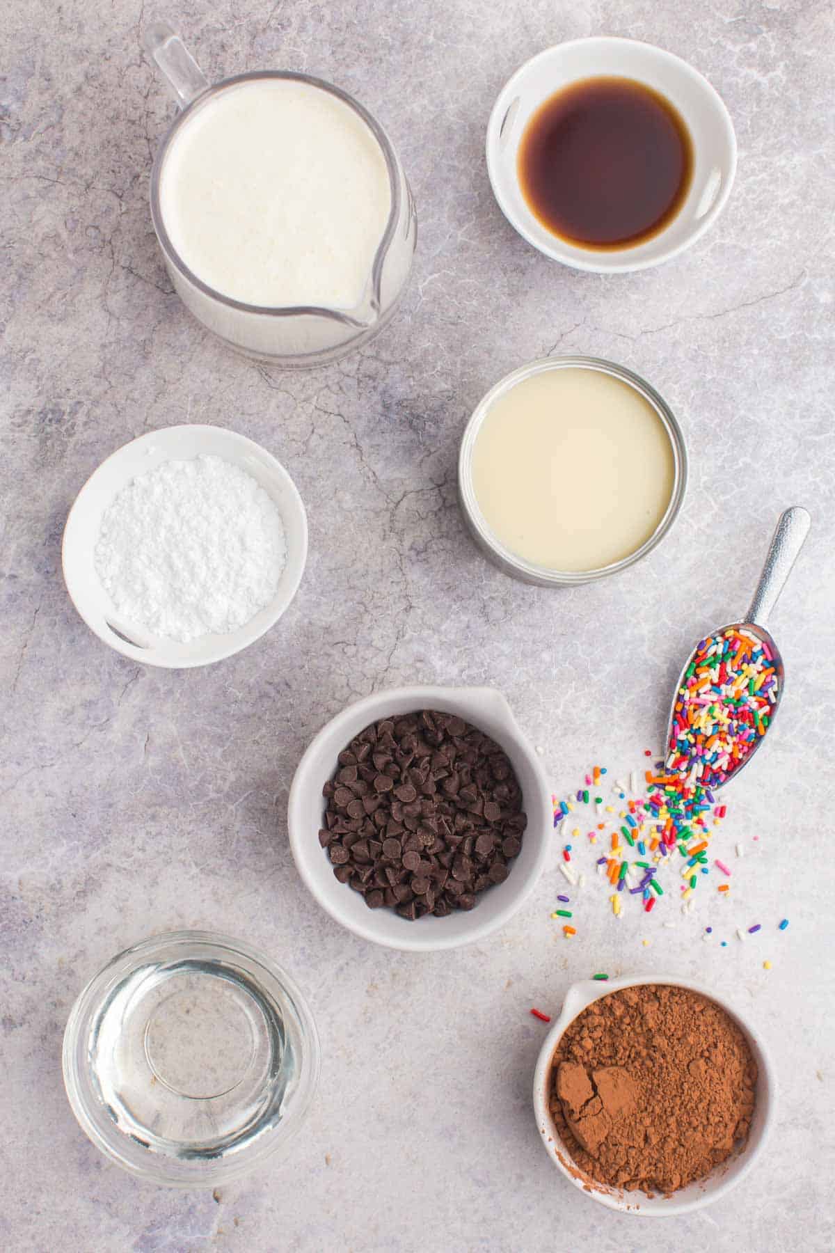 bowls of heavy cream, sweetened condensed milk, cocoa powder, vanilla extract, powdered sugar, corn syrup, chocolate chips, and rainbow sprinkles