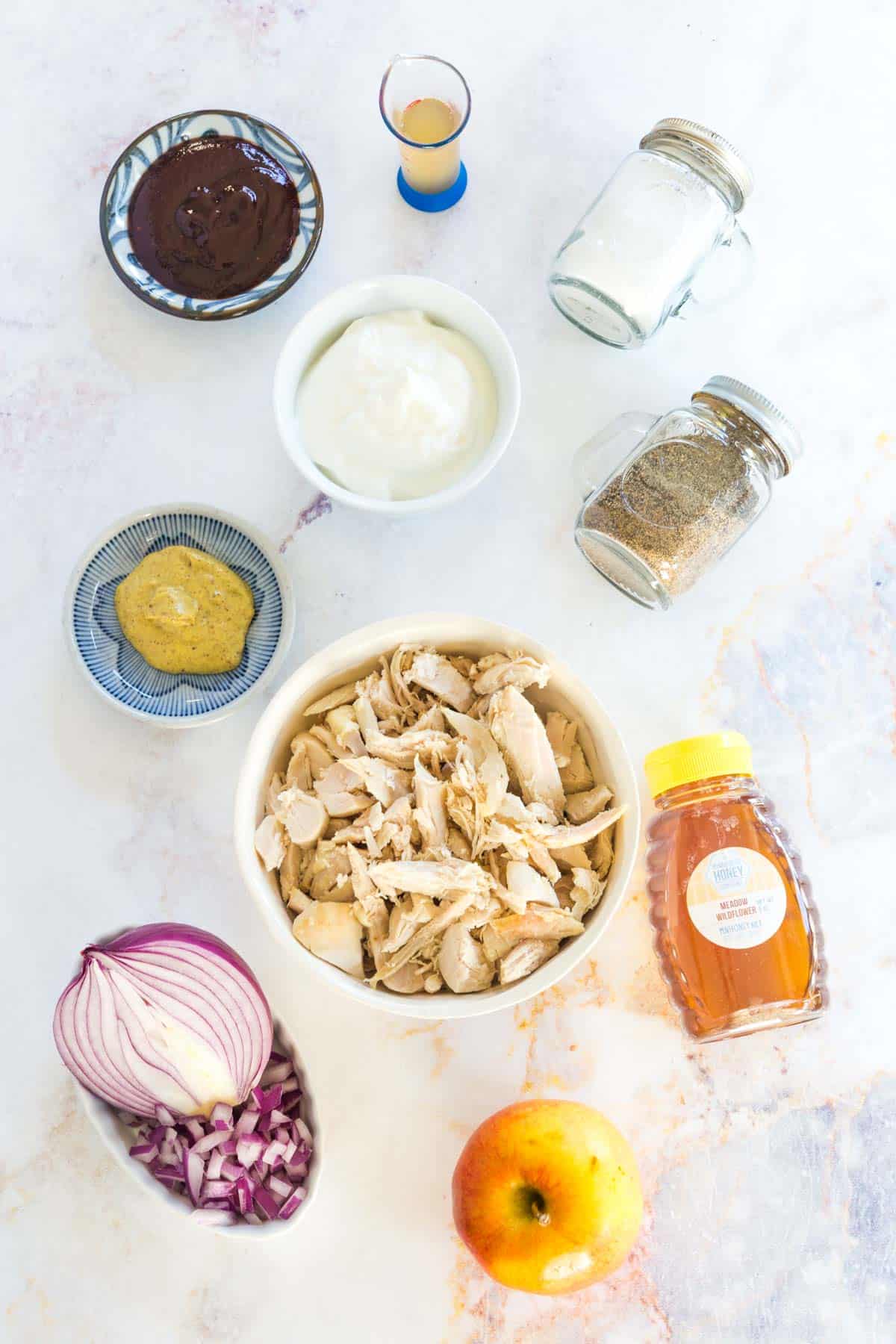 bowls of cooked chicken, Greek yogurt, mustard, barbecue sauce, apple cider vinegar, and red onion, an apple, a bottle of honey, and salt and pepper shakers