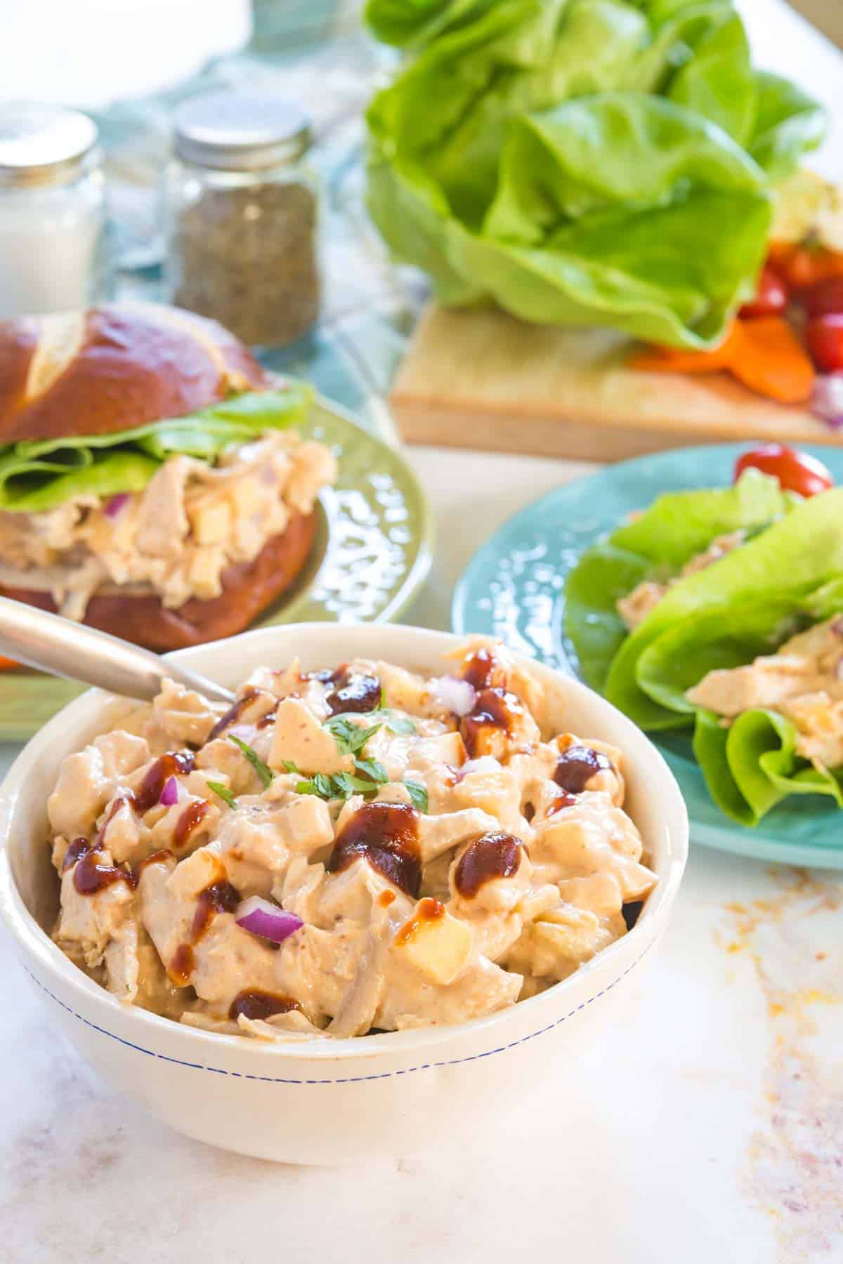 a white bowl filled with barbecue chicken salad drizzled with bbq sauce, and sandwiches and lettuce in the background
