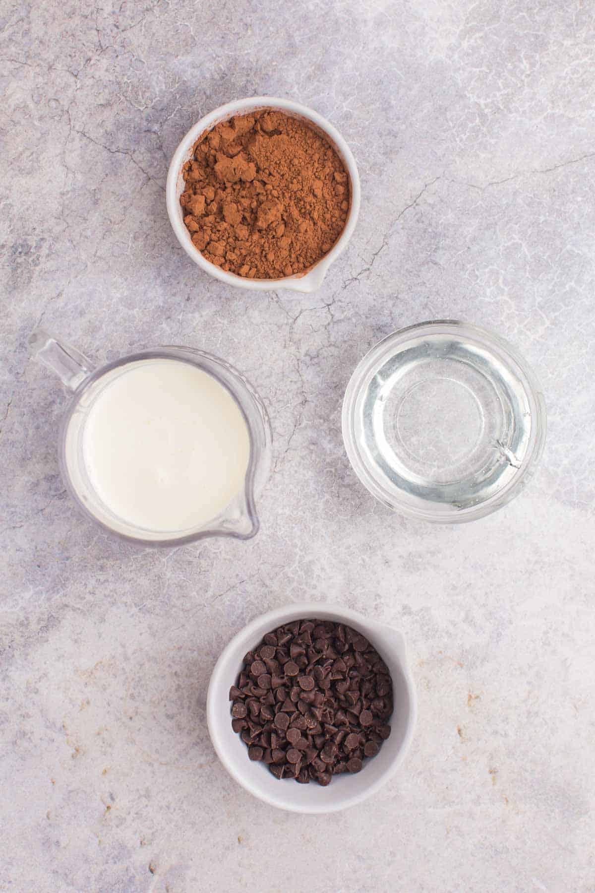 bowls of corn syrup, chocolate chips, and cocoa powder and a pitcher of heavy cream on a marble tabletop