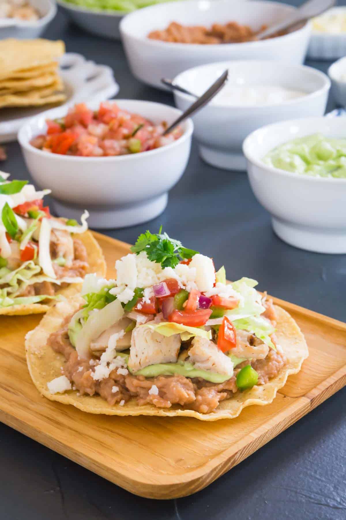 chicken tostada with all the toppings on a wooden serving platter with bowls of toppings behind it