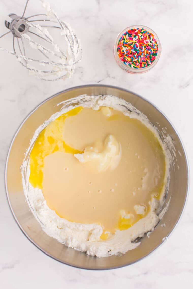 sweetened condensed milk and melted butter added to the bowl of whipped cream with a small bowl of rainbow sprinkled next to it