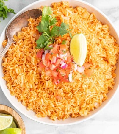 white bowl of spanish rice topped with chopped tomatoes and onions surrounded by bowls of salsa and limes