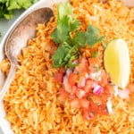 bowl of spanish rice with a serving spoon topped with chopped tomatoes, onions, and cilantro