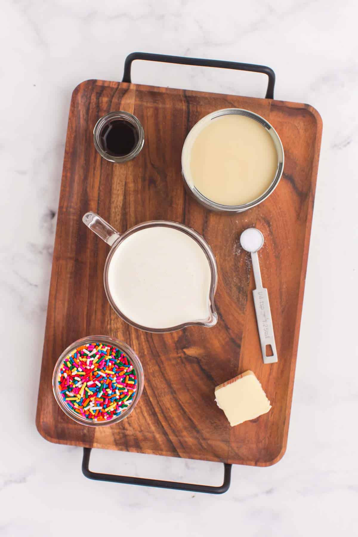 measuring cup of heavy cream, can of sweetened condensed milk, jar of vanilla extract, cube of butter, and a bowl of rainbow sprinkles on a wooden platter