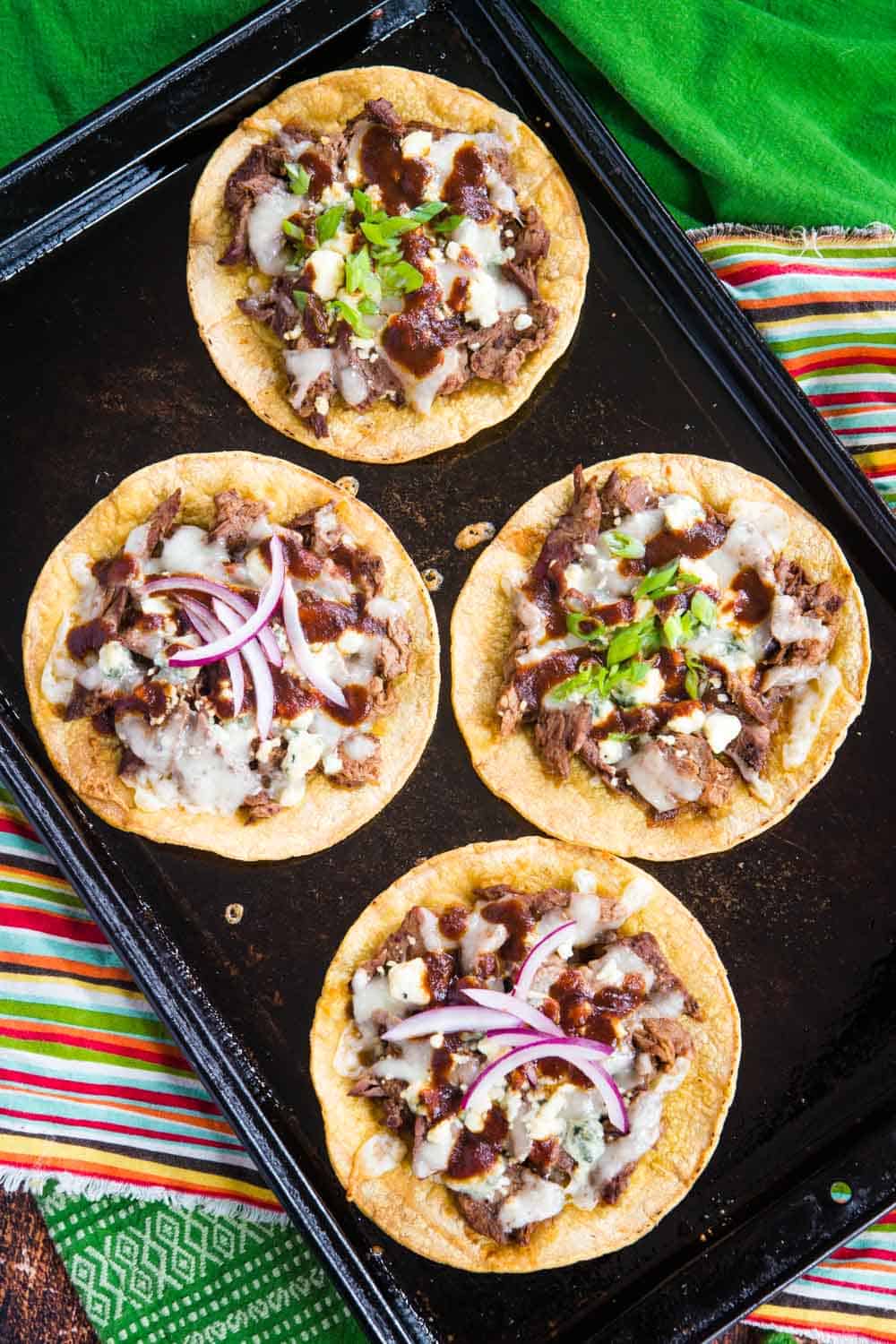 four steak tostadas on a baking sheet on top of green and multicolored striped napkins